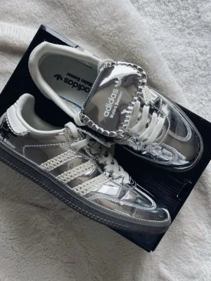 Adidas x Wales Bonner silvriga sneakers - Snygga sneakers från Adidas i samarbete med Wales Bonner. Skorna har en spegelblank silverfärg med vita detaljer och klassiska tre ränder på sidan. Ovandelen är i glansigt material med stickade inslag och bred plös med logga. Perfekta för dig som vill sticka ut.
