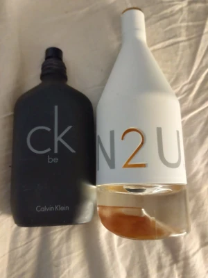 Calvin Klein parfymflaskor - Cirka 80% kvar i båda....Två parfymflaskor från Calvin Klein: en svart matt flaska med 'ck be' (Unisex) och en vit flaska med guld- och silvertext 'IN2U'.(Dam)...... Båda har modern design och tydliga logotyper. Perfekt för dig som gillar stilrena och trendiga dofter.