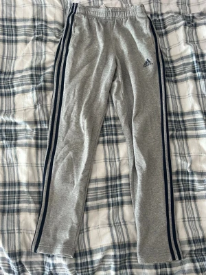 Grå Adidas mjukisbyxor med blå ränder - Grå mjukisbyxor från Adidas med blå ränder längs sidorna och elastisk midja. Byxorna har en klassisk rak passform och är tillverkade i mjukt material, perfekt för en avslappnad stil. Adidas-logga på vänster lår.