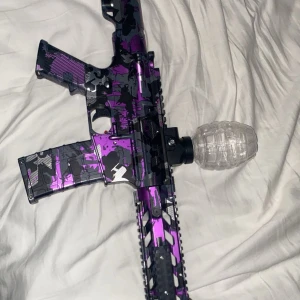 Lila och svart gelblaster gevär - Säljer en gelblaster i coolt lila, svart och grått camomönster. Geväret har en transparent behållare för gelkulor och detaljer i plast och metall. Perfekt för roliga utomhuslekar eller samling. Unik design som sticker ut bland vanliga leksaksvapen. Väldigt rolig och perfekt ifall du vill dominera vatten krig nu i sommar. Har använt den 3 gånger typ. CE märkt. Det ingår laddare. Hör av er vid minsta lilla fråga!!!!!