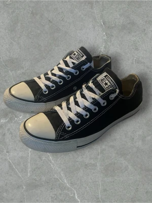 Svarta Converse All Star låga sneakers - Klassiska svarta Converse All Star sneakers med vit tå och vita snören. Skorna har lågt skaft, gummisula och ovandel i canvas. Ikonisk logga på plösen och snygga kontrastsömmar. Perfekta för en avslappnad och stilren look.