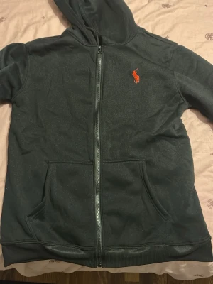Mörkgrå hoodie från Polo Ralph Lauren - Det är storlek L. Mörkgrå hoodie med dragkedja från Polo Ralph Lauren. Klassisk design med huva, kängurufickor och röd broderad logga på bröstet. Mjuk bomullsblandning som är skön att ha på sig. Perfekt för en avslappnad och stilren look.