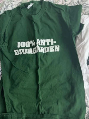 Hammarby t-shirt - Säljer en grön t-shirt med trycket '100% ANTI-DJURGÅRDEN' i vit text på bröstet. Klassisk passform med rund hals och korta ärmar. Perfekt för dig som vill visa ditt lagstöd på ett tydligt sätt.