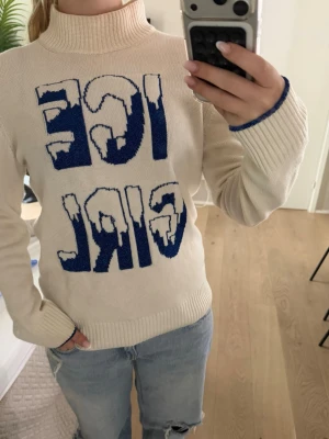 Vit stickad tröja MC2 Saint Barth - Vit stickad polotröja med blå texten 'ICE GIRL' framtill från MC2 Saint Barth. Tröjan har långa ärmar, ribbstickade muddar och hög krage. Materialet är mjukt och passar perfekt för kyliga dagar. Snygg kontrast med blå detaljer vid ärmslut.