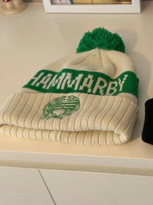 Hammarby mössor - Säljer två stickade mössor från Hammarby. Ena är vit med grön text, grön boll och grön tofs, andra är helsvart med liten Hammarby-flagga framtill. Perfekta för dig som vill visa ditt stöd för laget och hålla dig varm.