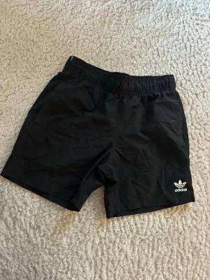 Svarta badshorts från Adidas Originals - Svarta badshorts från Adidas Originals med elastisk midja och klassisk logga framtill. Shortsen har en enkel design med fickor på sidorna och en bakficka. Inga skador eller fel med shortsen. storlek S