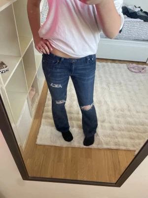 Mörkblå bootcut-jeans från AG - Säljer ett par mörkblå bootcut-jeans från AG med slitna detaljer och hål på knäna. Jeansen har klassisk femficksdesign, låg midja och är tillverkade i stretchig denim för en bekväm passform. Passar som S/XS