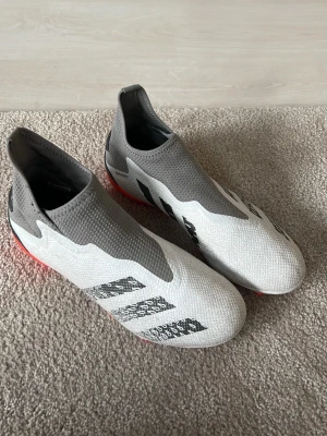 Adidas vita och grå fotbollsskor - Snygga fotbollsskor från Adidas i vitt och grått med röda dobbar. Skorna har en strumpliknande passform utan snörning och är tillverkade i syntetmaterial för bästa grepp och känsla på planen. Perfekta för dig som vill sticka ut på matchen.