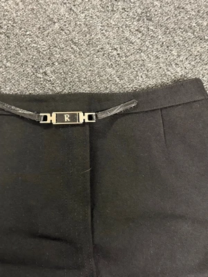 Svarta kostymbyxor med metallspänne - Stilrena svarta kostymbyxor med ett snyggt metallspänne framtill där bokstaven R syns. Byxorna har raka ben och är tillverkade i ett mjukt tyg som ger en klassisk look. Perfekta för dig som vill ha en clean och modern stil.
