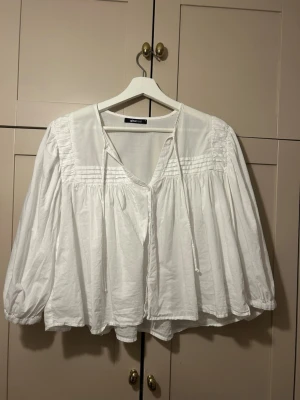 vit blus från Gina Tricot -  vit blus från Gina Tricot med v-ringning och knytband vid halsen. Blusen har veckade detaljer upptill och vida, långa ärmar med resår vid ärmslut🤍