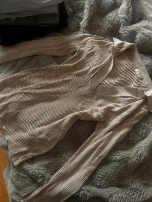 Beige ribbad blus med krage - Stilren beige ribbad blus med lång ärm och klassisk krage. Djup v-ringning framtill och mjukt, stretchigt material som sitter snyggt på kroppen. Perfekt att styla både upp och ned 