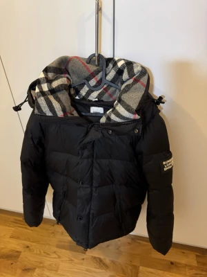 Svart dunjacka från Burberry - Snygg svart dunjacka från Burberry med klassiskt rutigt foder i grått, svart, vitt och rött. Jackan har dragkedja, tryckknappar och justerbar huva. Stor logotyp på ärmen. Perfekt för kalla vinterdagar.