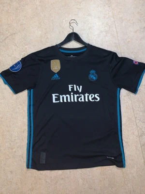 L. Riktig Real Madrid tröja 2017/2018  - Använt den max 6 gånger vill sälja den för har börjat gymma vill jätte gärna köpa gym hörlurar Säljer en svart Real Madrid matchtröja från Adidas med Ronaldo 7 på ryggen. Tröjan har blå detaljer, klubbmärke, Champions League-patchar och Fly Emirates-tryck på bröstet. Perfekt för fotbollsfans som vill sticka ut på planen eller läktaren.