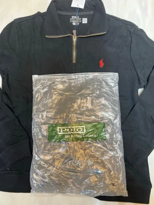 Svart tröja med dragkedja från Polo Ralph Lauren - Svart långärmad tröja från Polo Ralph Lauren med halv dragkedja och klassisk röd broderad logga på bröstet. Tröjan har hög krage och är tillverkad i mjukt material, perfekt för en stilren och avslappnad look.