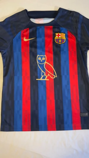 Barcelona x Drake matchtröja Nike - Snygg Barcelona matchtröja i blått och rött med Drake-tryck och nummer 50 på ryggen. Framtill finns Nike-logga, Barca-emblem och OVO-uggla i guld. Tillverkad i lätt och ventilerande material, perfekt för fotboll eller streetwear.