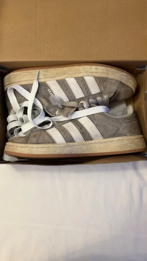 Grå Adidas Campus sneakers i mocka - Gråa Adidas Campus sneakers i mocka med klassiska vita ränder på sidorna och vit logga på plösen. Skorna har vita, breda skosnören och en beige sula i gummi. Perfekta för en avslappnad streetstyle-look.