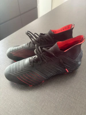 Adidas Predator svarta fotbollsskor - Säljer ett par svarta Adidas Predator fotbollsskor med röda detaljer och hög strumpliknande krage. Skorna har snörning, mönstrad ovandel i syntetmaterial och texten 'Primeknit' på insidan. Perfekta för dig som vill ha grym kontroll på planen.