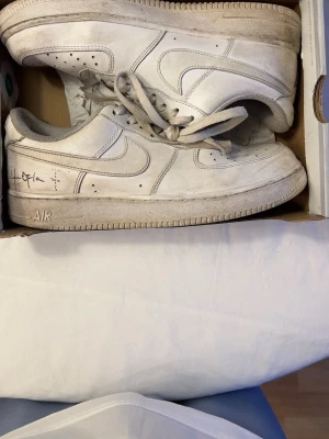 Vita Nike Air Force 1 sneakers - Klassiska vita Nike Air Force 1 sneakers i läder med grå detaljer och perforerad tå för extra ventilation. Swoosh-logga på sidorna och AIR-märkning på sulan. Lågt skaft och snörning framtill. Perfekta för en streetwear-look.