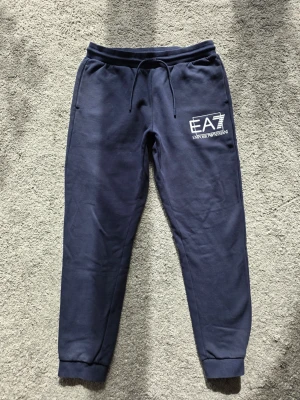 EA7 Emporio Armani till ditt pris!!! - Säljer ett par mörkblå mjukisbyxor från EA7 Emporio Armani med dragsko i midjan och ribbade muddar vid bensluten. Byxorna har en snygg vit logga på vänster lår och är tillverkade i mjukt material som känns riktigt skönt mot huden.