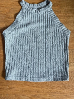 Grå ribbad ärmlös topp från Bershka - Säljer en grå ribbad topp från Bershka. Toppen är ärmlös med hög hals och har en tight passform. Perfekt att styla med jeans eller kjol för en trendig look.