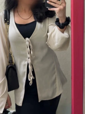 Beige satinblus med knytning - Beige blus i satinliknande material med djup V-ringning och två knytband framtill. Blusen har långa, lätt utsvängda ärmar och en avslappnad passform. Inga skador eller defekter. 