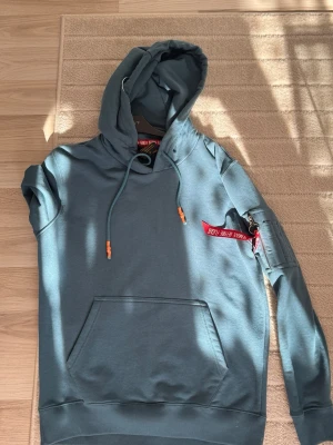 Blå hoodie från Alpha Industries - Snygg blå hoodie från Alpha Industries med klassisk känguruficka och huva med dragsko. På ärmen sitter en röd tag med texten 'Remove Before Flight' som ger en cool detalj. Perfekt för dig som gillar streetwear och vill sticka ut lite extra.