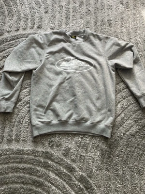 Grå sweatshirt med tryck - Snygg grå sweatshirt från Fruit of the Loom med ett vitt tryck av ett fartyg i oval ram på bröstet. Tröjan har rund hals, långa ärmar och ribbade muddar vid ärmslut och nederkant. Perfekt för en avslappnad och stilren look.