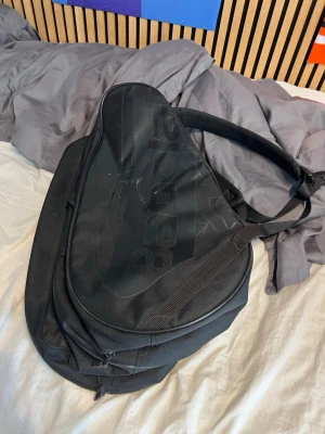 Svart ryggsäck från Babolat - Snygg svart ryggsäck från Babolat med sportig design och diskret logga på framsidan. Väskan har en unik form, perfekt för racketsport, och en gul dragkedja som sticker ut. Tillverkad i slitstarkt material och har både bärhandtag och axelrem.