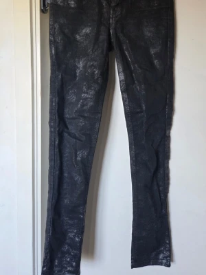 Svarta jeans från Lee med lite metallicfärg i mörkt silver goth rock metal - Säljer ett par svarta jeans från Lee med glansig/silvrig finish och smal passform. Jeansen har klassisk femficksdesign och knappgylf. Materialet är denim med en metallic-liknande yta som ger en cool look. Stl saknas, uppskattas till XXS/XS. Midjemått: ca 62 cm. Innerbenslängd: ca 70 cm. 