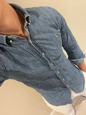 Blå jeansskjorta från Ralph Lauren - Säljer en klassisk blå jeansskjorta från Ralph Lauren med knappstängning och broderad logga på bröstet. Skjortan har lång ärm och normal passform, perfekt till jeans eller chinos för en avslappnad look.
