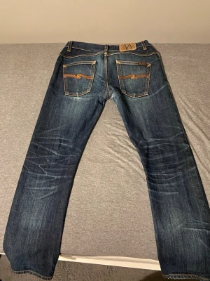 Nudie Jeans (Selvage) - Säljer ett par mörkblå jeans från Nudie Jeans med klassisk femficksdesign och snygga kontrastsömmar. Bakfickorna har Nudies ikoniska broderi i orange. Jeansen är raka i modellen och tillverkade i denim av bomull. (Selvage)