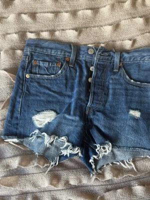 Blå Levi's jeansshorts med slitningar - Storlek W 26. Säljer ett par blå jeansshorts från Levi's med råa, fransiga kanter och slitningar på framsidan. Knappar framtill