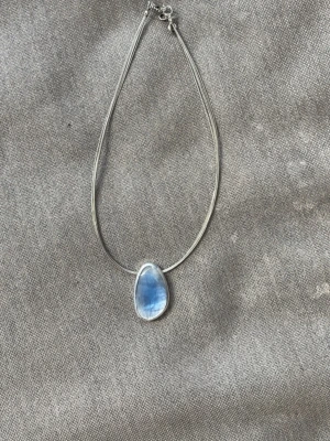Silverhalsband med blå sten, trasigt ” längdjustrringskejda”  - Snyggt halsband i silver med en droppformad blå sten som hänger i mitten. Kedjan är tunn och elegant, och stenen har en skimrande effekt som gör att den verkligen står ut. Perfekt för dig som gillar stilrena och moderna smycken.