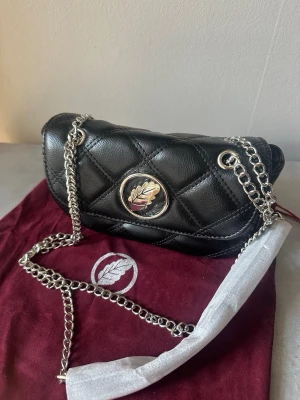 Svart  handväska  - Cool svart handväska  med silverfärgad kedja och ett snyggt lövmärke på framsidan från ioaku. Perfekt för dig som gillar stilrena accessoarer.