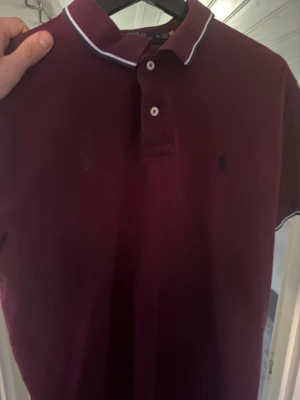 Vinröd polotopp från Ralph Lauren - Säljer en vinröd polotopp från Ralph Lauren med klassisk krage och två knappar framtill. T-shirten har vit kant på kragen och är kortärmad. Custom slim fit-modell, perfekt för dig som gillar en snygg och stilren look.