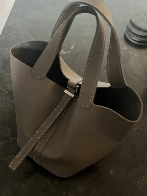 H - väska taupe - H - väska i en grå brun nyans. Medföljer axelrem och neccessär. 