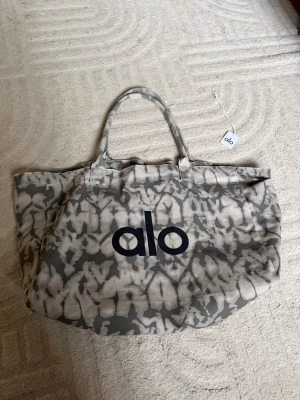 Mönstrad väska från Alo - Cool väska från Alo med grått och vitt abstrakt mönster. Väskan är rymlig med två handtag och har Alo-loggan tryckt i svart på framsidan. Tillverkad i kraftig canvas, perfekt för att bära allt du behöver ☺️🤍