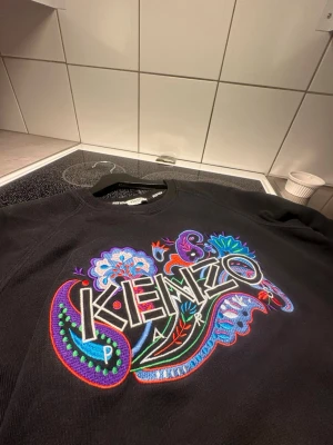 Svart Kenzo sweatshirt med brodyr - Cool svart sweatshirt från Kenzo med färgglad broderad logga och blommönster på bröstet. Tröjan har rund halsringning och långa ärmar. Perfekt för dig som vill sticka ut med en unik och trendig design.