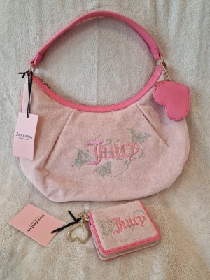 Juicy Couture rosa axelväska - Söt axelväska i ljusrosa mocka med rosa detaljer från Juicy Couture. Väskan har broderad Juicy-logga, hjärtformad nyckelring och matchande liten plånbok. Perfekt för dig som gillar en trendig och feminin look.