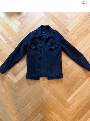 Svart jeansjacka från Zara - Stilren svart jeansjacka från Zara med klassisk krage, knäppning framtill och två bröstfickor med lock. Jackan har raka ärmar och en tidlös design som passar till många olika outfits.