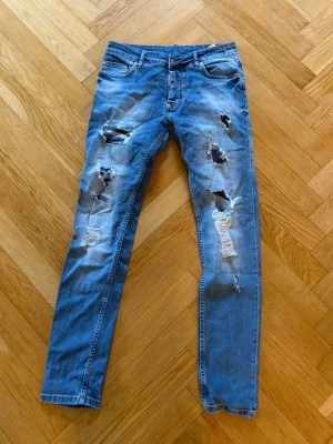 Blå slitna dsq jeans  - Säljer ett par blå  jeans med flera stora slitningar och hål på  framsidan Jeansen har klassisk femficksdesign Perfekta för en avslappnad och trendig streetstil.