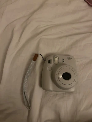 Fujifilm Instax Mini 9 med rosa fodral - Fujifilm Instax Mini 9 är en vit, kompakt direktfilmskamera i plast. Den har en rundad design och kommer med ett handledsband samt ett rosa fodral med axelrem. Perfekt för att ta instantbilder och ha med sig på äventyr.