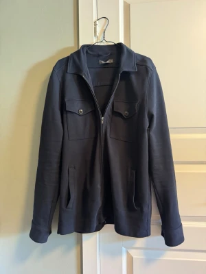 Blå overshirt från Dressmann - Stilren svart overshirt från Dressmann med dragkedja framtill, två bröstfickor med lock och knappar samt två sidofickor. Overshirten har klassisk krage och är tillverkad i ett mjukt, strukturerat tyg som ger en avslappnad look.