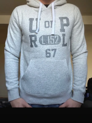 Ljusgrå hoodie med tryck och huva - Ljusgrå hoodie från U.S. Polo Assn. med stort tryck framtill i mörkgrått. Mjuk bomullsblandning och ribbade muddar vid ärmslut och nederkant. Perfekt för en avslappnad stil✅ Tveka inte på att fråga vid behov😁