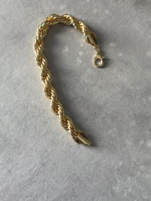 10mm Tjockt cordell guldarmband - Snyggt armband i guldfärgad metall med tvinnad design och robust karbinlås. Armbandet har en chunky känsla och passar perfekt för att ge din outfit lite extra bling. Längden är cirka 21 cm.