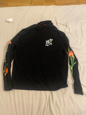 Svart HUGO tröja med blommor - Cool svart långärmad tröja från HUGO med stora vita bokstäver och orange blommor på ryggen samt ärmarna. Framtill finns en vit blom-illustration på bröstet. Perfekt för dig som gillar unika prints och streetstyle.