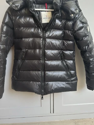 Svart dunjacka från Moncler - Snygg svart dunjacka från Moncler med glansig finish, huva och två dragkedjeförsedda fickor framtill. Jackan har quiltad design och Moncler-logga på ärmen. Perfekt för kalla vinterdagar och riktigt trendig look.