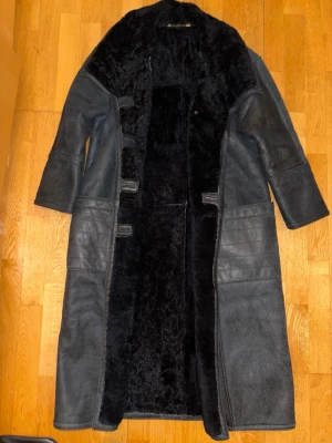 äkta fårskinnskappa – Hollies – svart shearling, lång modell - Äkta fårskinnskappa från Hollies i klassisk lång modell. Unisex, passar både dam och herr beroende på önskad passform.  Tillverkad i äkta shearling/fårskinn med varm och mjuk päls på insidan. Kraftigt, gediget ytterplagg av hög kvalitet som håller i många år. Tidlös design som fungerar lika bra till vardag som till mer uppklädda tillfällen.  Färgen är svart/mörkgrå med naturlig patina som är typisk för äkta skinn och ger karaktär. Kappan är begagnad men i bra skick för sin ålder.