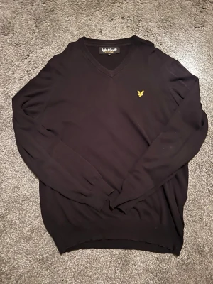 Svart V-ringad tröja Lyle & Scott - Svart långärmad tröja från Lyle & Scott med V-ringning och klassisk gul logga på bröstet. Tröjan har ribbade muddar vid ärmslut och nederkant. Perfekt för en stilren och enkel look.