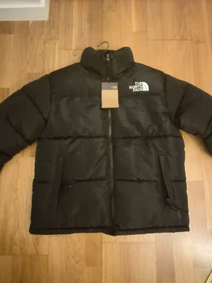 Svart pufferjacka från The North Face - Säljer en svart OBS Replica pufferjacka från The North Face med hög krage och dragkedja framtill. Jackan har två stora fickor på sidorna och en på insidan och logga broderad i vitt på bröstet och rygen . Klassisk boxig passform och tjockt vadderad för extra värme. Perfekt för kalla vinterdagar.